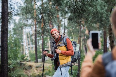 blurred woman taking photo of cheerful boyfriend on smartphone with blank screen 