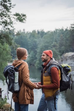 young and joyful couple holding hands while looking at each other near lake in forest 