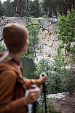 back view of blurred woman standing with hiking sticks near cliff and lake
