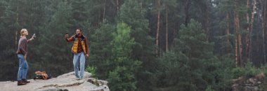 happy couple with smartphones standing on rocky cliff during rain, banner