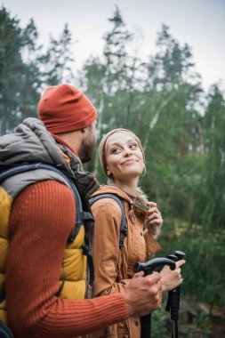 happy young couple with backpacks trekking together with hiking sticks and looking at each other 