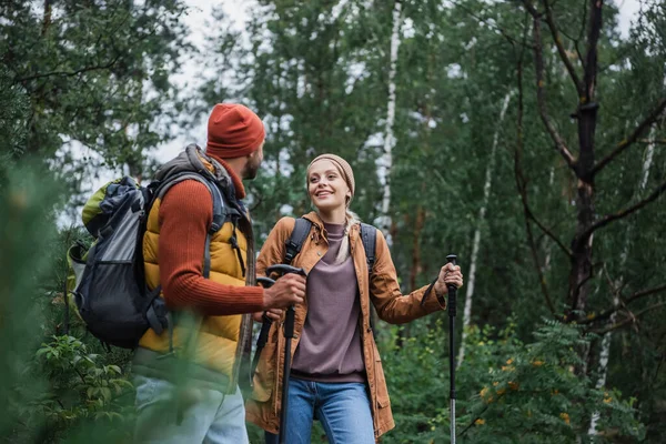 happy woman with hiking sticks walking and looking at boyfriend in forest