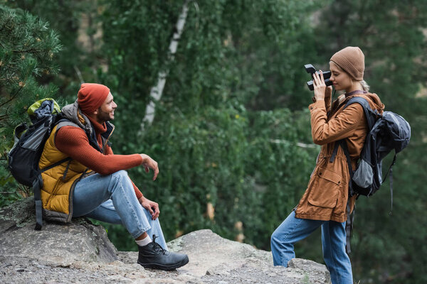 woman with backpack taking photo of happy boyfriend on vintage camera in forest 