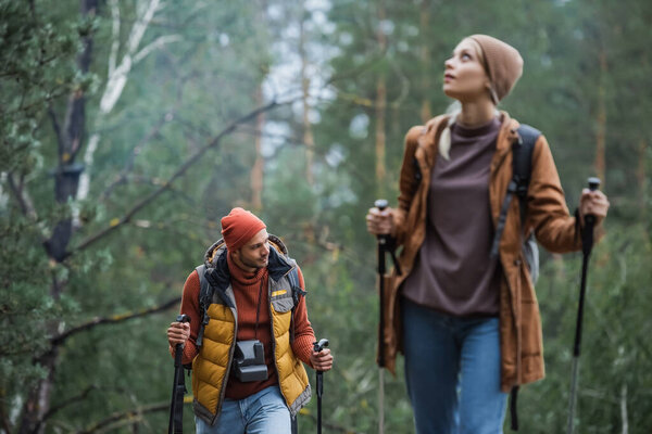 man with vintage camera looking away near woman holding hiking sticks on blurred foreground 