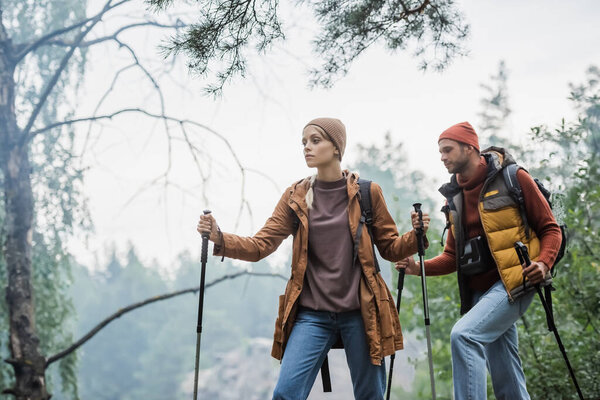 couple in hats holding hiking sticks while trekking in forest