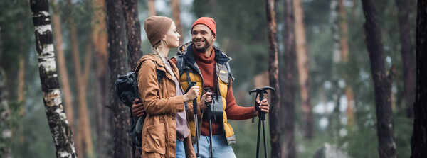 cheerful couple in hats holding hiking sticks while hugging in forest, banner