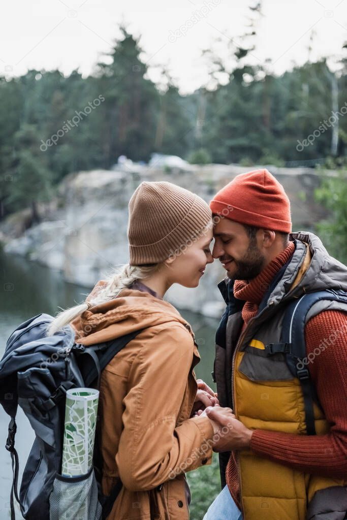 young and joyful couple with closed eyes holding hands near lake in forest 