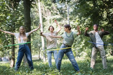 Cheerful multiethnic teenagers twisting hula hoops on grass  clipart
