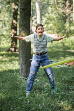 Excited teen girl twisting hula hoop on grass in park  clipart