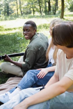 Gülümseyen Afrikalı Amerikalı genç parkta bulanık arkadaşlarının yanında akustik gitar çalıyor. 