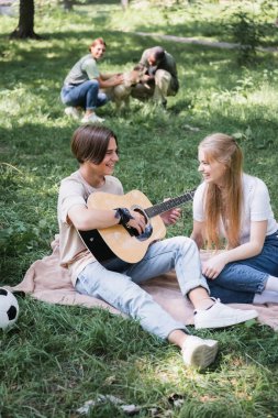 Gülümseyen gençler çimenlikte köpekli bulanık arkadaşlarının yanında akustik gitar çalıyorlar. 