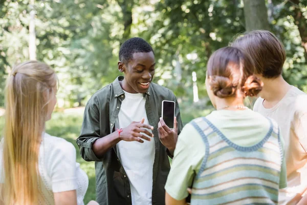 Gülümseyen Afrikalı Amerikalı Genç Parkta Bulanık Arkadaşlarına Akıllı Telefon Gösteriyor — Stok fotoğraf