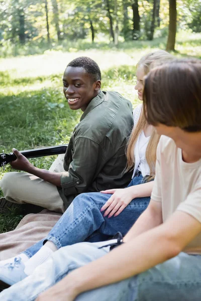 Sonriente Adolescente Afroamericano Tocando Guitarra Acústica Cerca Amigos Borrosos Parque —  Fotos de Stock