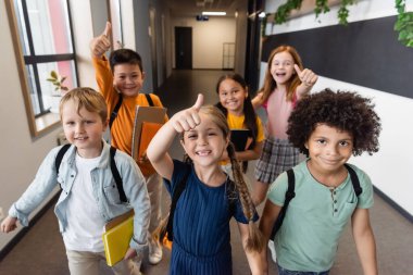 Neşeli çok kültürlü okul çocukları okul koridorunda baş parmak gösteriyorlar.