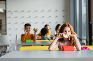 sad redhead girl eating sandwich near schoolkids on blurred background clipart