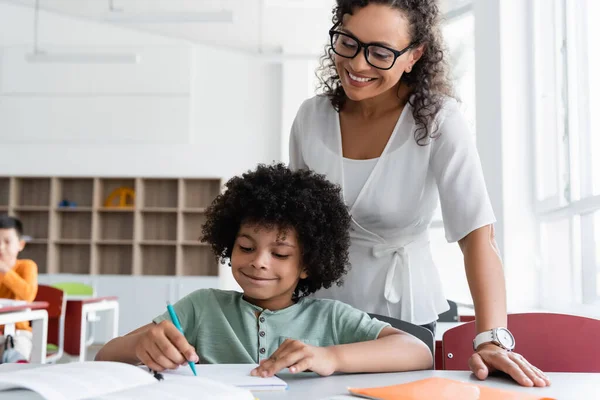 Glimlachende Leraar Kijken Naar Afrikaanse Amerikaanse Jongen Schrijven Tijdens Les — Stockfoto