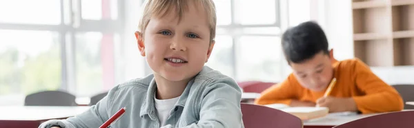 Vreugdevolle Jongen Kijken Naar Camera Terwijl Aziatische Klasgenoot Schrijven Wazig — Stockfoto