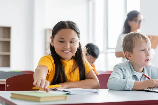 Cheerful Asian Schoolgirl Reaching Book While Sitting Desk Classmate — Stock Photo, Image