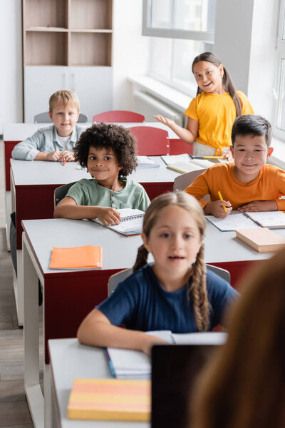 smiling asian schoolgirl talking during lesson near multicultural classmates