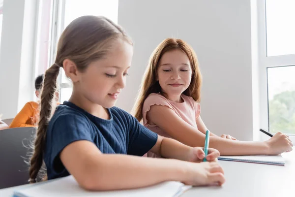 Schoolmeisje Schrijven Notebook Buurt Glimlachende Klasgenoot Tijdens Les — Stockfoto