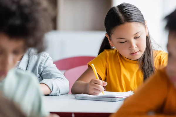 Enfocado Asiático Chica Escritura Durante Lección Cerca Borrosa Multiétnico Colegiales —  Fotos de Stock