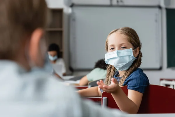 schoolgirl in medical mask pointing with hand near blurred boy during lesson