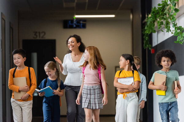 smiling african american teacher pointing with hand while talking to multiethnic children in school hall