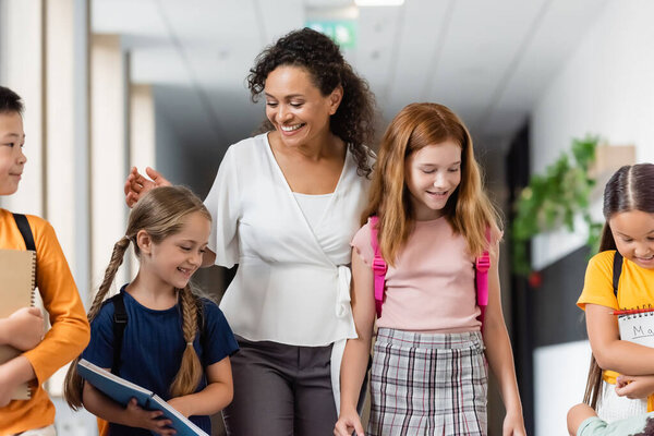 happy african american teacher walking with multiethnic kids in school corridor