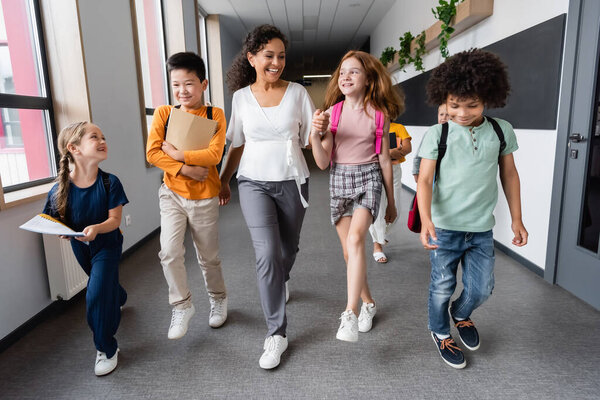 happy african american teacher holding hands with girl near multiethnic pupils in school hall