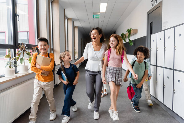 laughing african american teacher walking with multiethnic kids in school hall