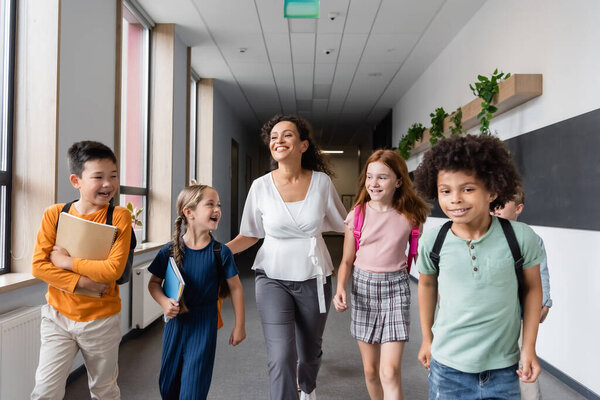 cheerful multicultural kids walking in school corridor with african american teacher