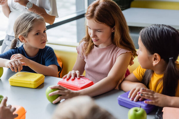 redhead girl holding apple and opening lunch box near multiethnic classmates