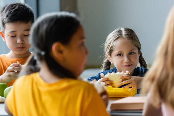 schoolgirl eating sandwich near blurred asian friends 