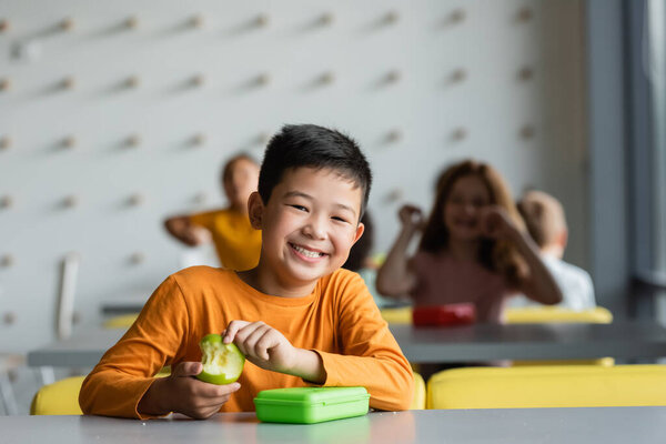 happy asian kid holding with fresh apple smiling at camera near blurred children in school canteen