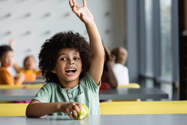 african american schoolboy waving hand near apple and kids on blurred background