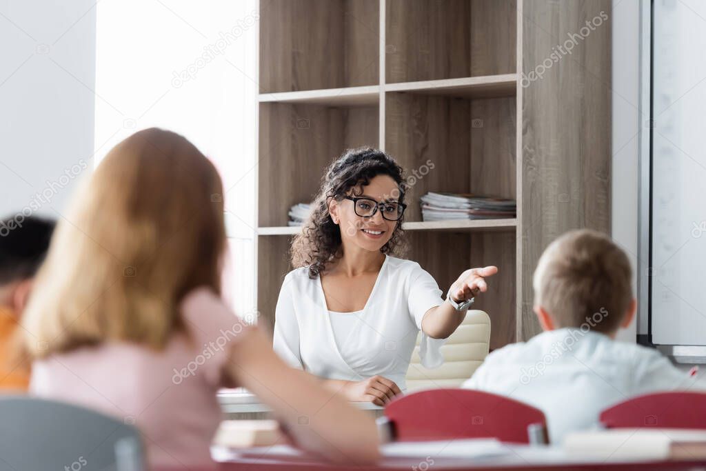 positive african american teacher pointing with hand near blurred pupils