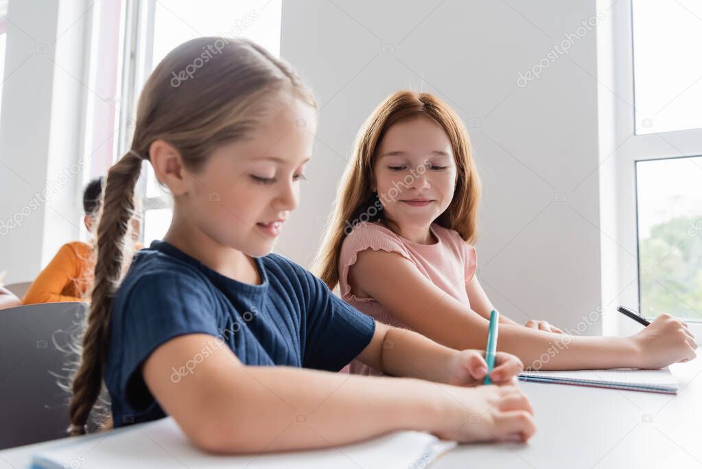schoolgirl writing in notebook near smiling classmate during lesson