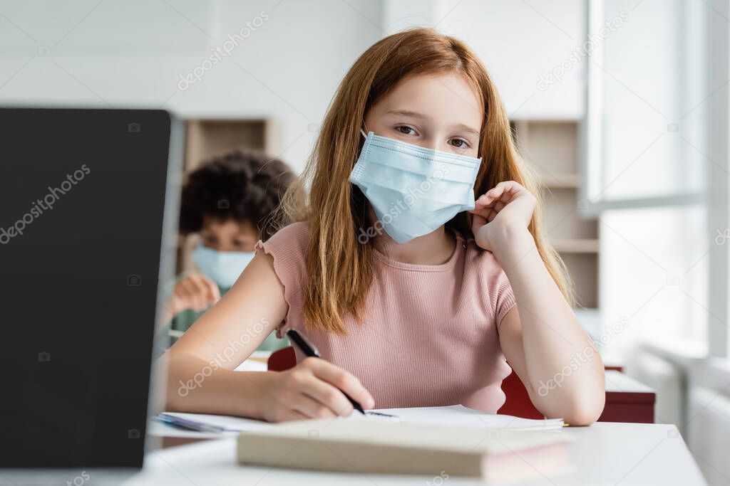 redhead girl in medical mask looking at camera during lesson