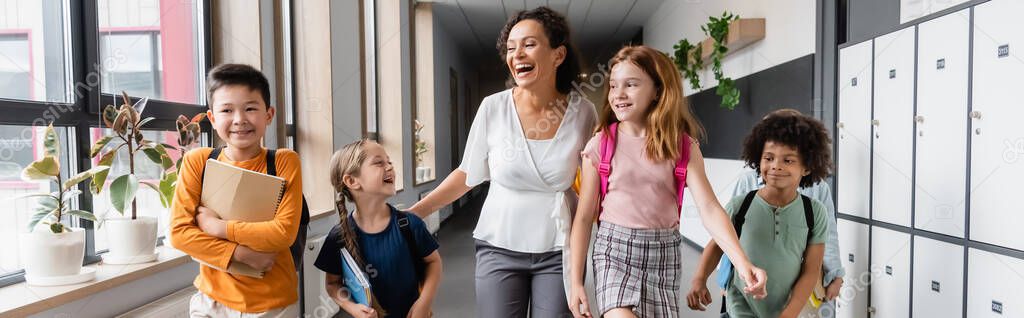 excited african american teacher laughing near multiethnic pupils in school hall, banner