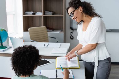 Gülümseyen öğretmen ders sırasında Afrikalı Amerikalı çocuğun yanındaki kitaba işaret etti.