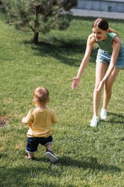 Neşeli bir anne, uzanmış elleri çimlerin üzerinde duruyor ve yeni yürümeye başlayan oğluna uzanıyor. 
