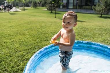 Yüzme şortlu çocuk plastik oyuncakla şişme havuzda duruyor.
