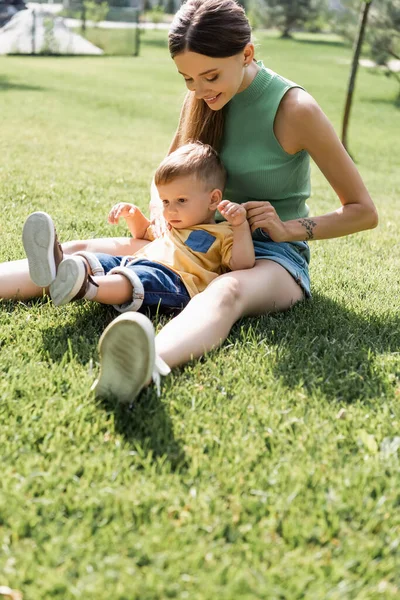 明るいお母さんは外で幼い息子と草の上に座って — ストック写真