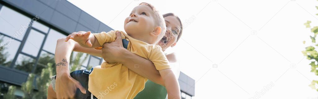 low angle view of amazed young mother holding in arms smiling toddler son, banner
