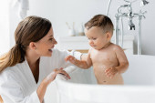 cheerful mother in bathrobe bathing wet toddler son in bathtub 