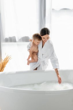 Bornozlu şefkatli anne kucağında çıplak bebek oğlu banyo köpüğüyle küvetin yanında. 