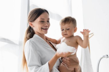 Bornozlu gülümseyen anne çıplak çocuğun yanında banyo köpüğü tutuyor. 
