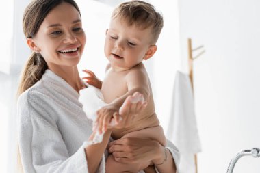 Bornozlu gülümseyen anne banyoda banyo köpüğü ve çıplak bir çocuk tutuyor.