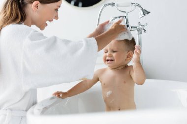 Bornozlu neşeli anne küvette banyo yapan küçük çocuk. 