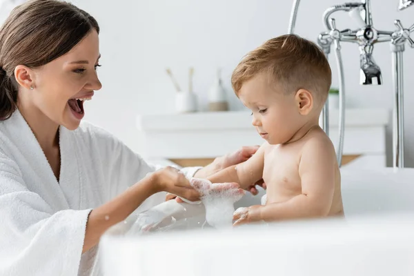 快乐的妈妈穿着浴衣 手里拿着泡沫浴在浴缸里蹒跚学步的男孩身边 — 图库照片